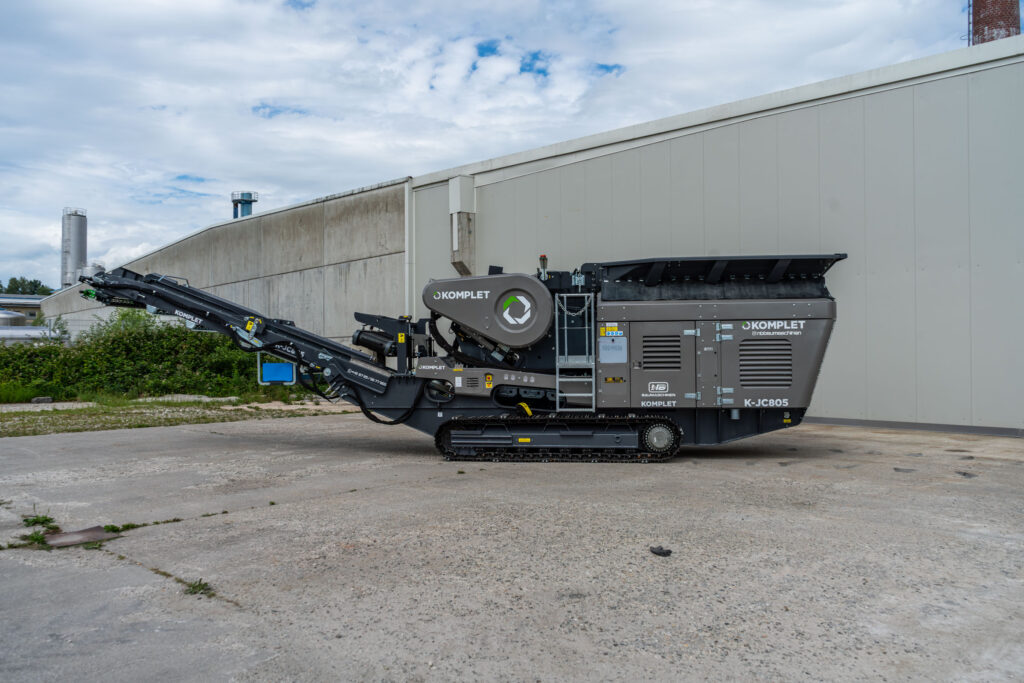 Schwarzer Komplet K-JC805 Backenbrecher mit Raupenfahrwerk und langem Förderband steht vor einer hellen Industriehalle unter bewölktem Himmel im Freien.
