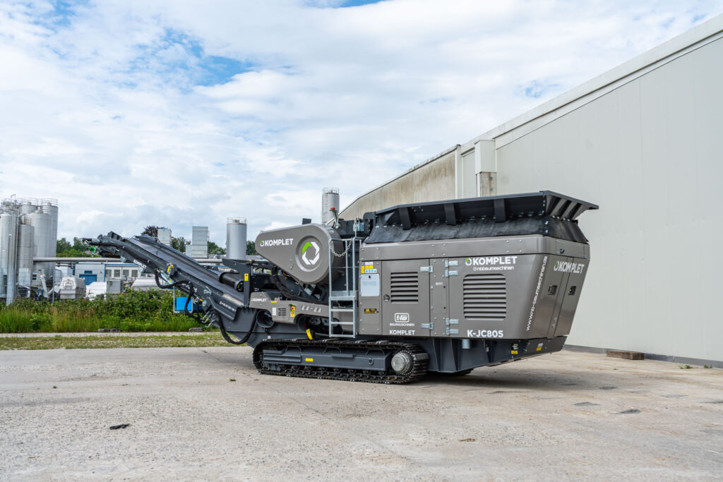 Der graue Komplet K-JC805 steht auf Raupenfahrwerk vor einer Industriehalle, mit langem Förderband, Einfülltrichter und NB-Baumaschinen-Branding.