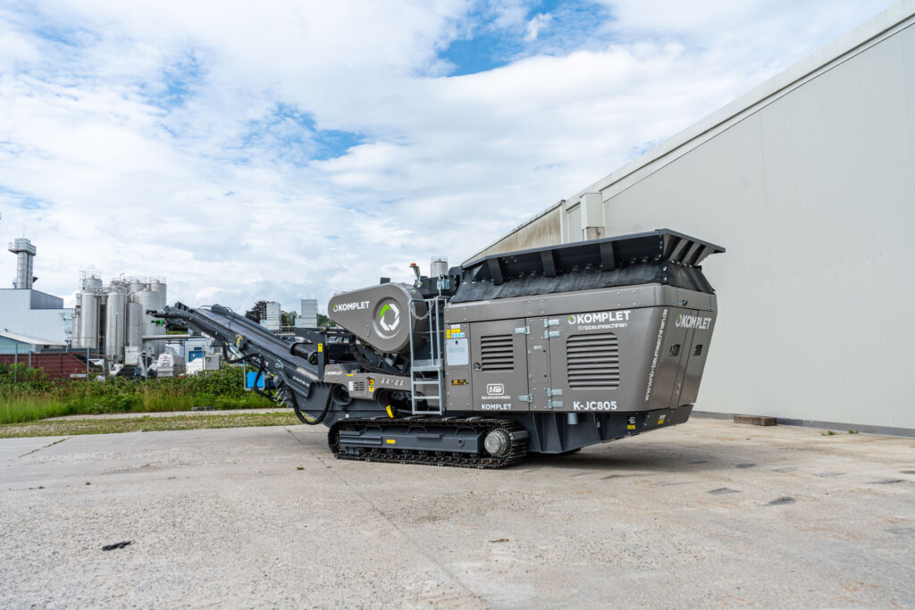 Ein grauer K-JC805 von Komplet steht auf Raupenfahrwerk vor einer Industriehalle; das lange Förderband ragt nach links unter wolkigem Himmel.