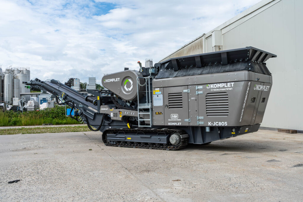 Grauer Komplet K-JC805 Raupen-Backenbrecher von NB Baumaschinen steht vor einer Industriehalle, mit Förderband, Trichter und Raupenfahrwerk.