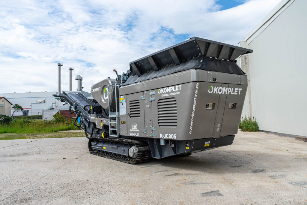 Der graue KOMPLET K-JC805 Backenbrecher steht auf Raupenfahrwerk vor einer Industriehalle, mit schwarzem Einfülltrichter und sichtbaren NB-Baumaschinen-Logos.