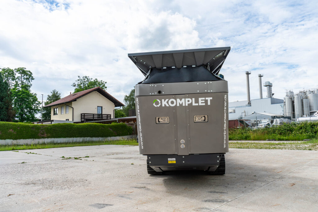 Großer grauer Komplet-Backenbrecher mit breitem Einfülltrichter steht auf Raupenfahrwerk vor Industriegebäuden und unter einem hellen, wolkigen Himmel.