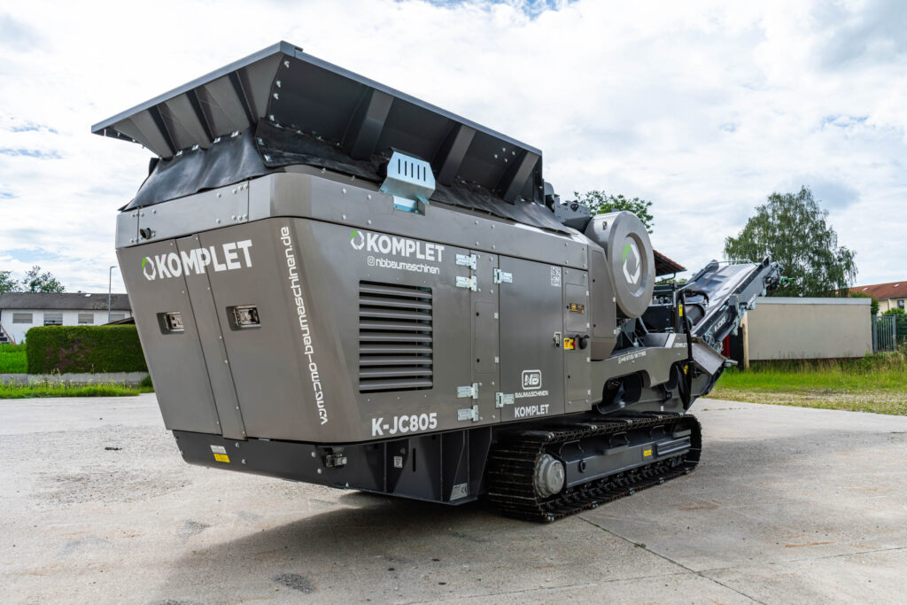 Der graue Komplet K-JC805 Backenbrecher steht auf Raupenfahrwerk unter bewölktem Himmel, mit großem Aufgabetrichter, Wartungsklappen und Förderband.