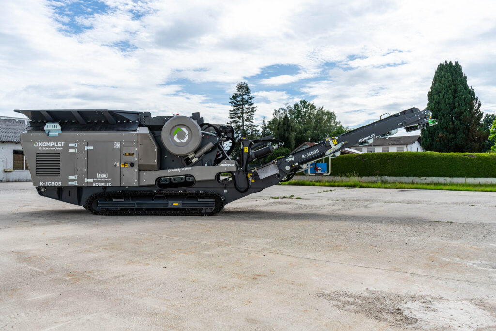 Der dunkelgraue Backenbrecher Komplet K-JC805 steht auf Raupenfahrwerk auf einem Betonplatz, mit ausgefahrenem Förderband unter wolkigem Himmel.