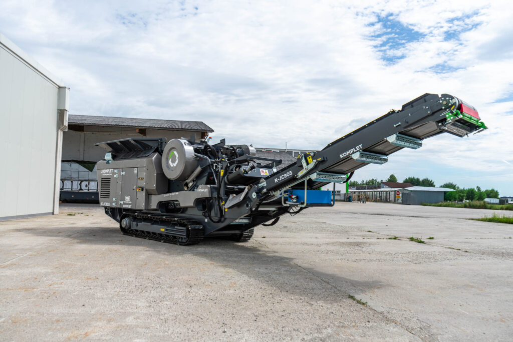 Der grauschwarze Komplet K-JC805 Raupen-Backenbrecher steht auf einem Betonhof, der Förderer ist ausgefahren, darüber liegt ein heller Himmel.