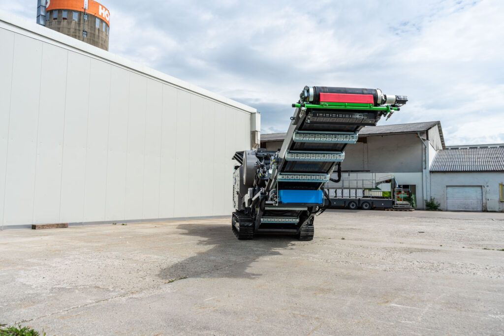 Ein mobiler Backenbrecher K-JC805 steht auf Raupenfahrwerk vor Industriegebäuden, mit hochgestelltem Förderband, grün-roten Bauteilen und breitem Arbeitsbereich.