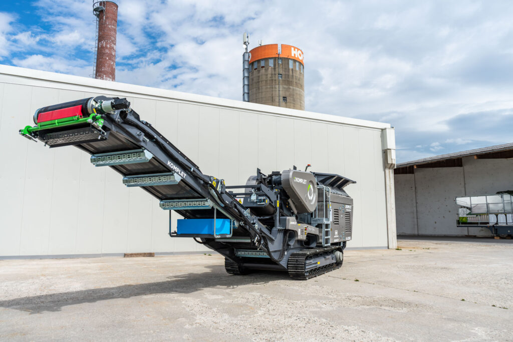 Ein schwarzer Komplet K-JC805 Brecher steht auf Raupenfahrwerk vor einer hellen Halle; das Förderband ist ausgefahren, dahinter ragen Silos auf.