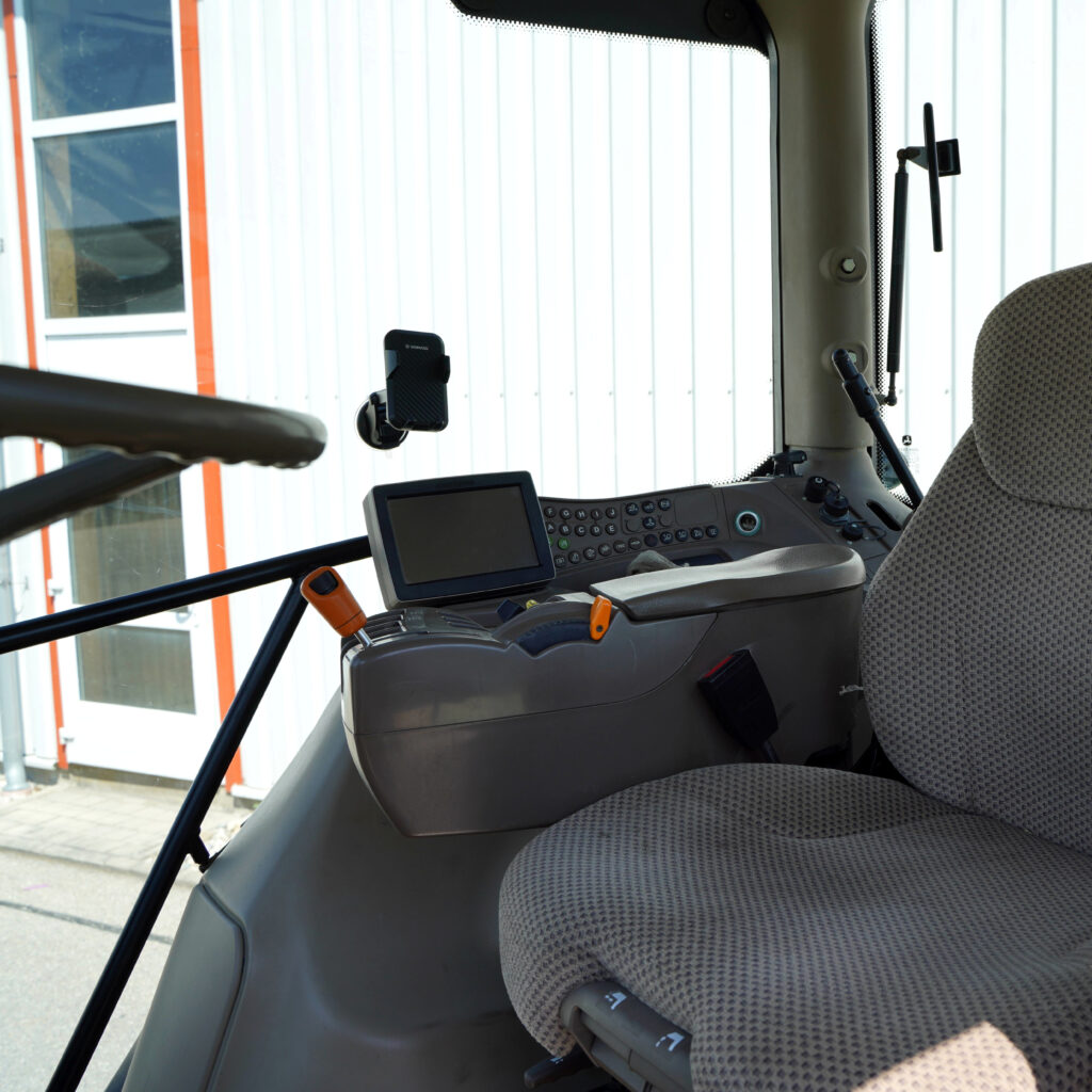 Graue Fahrerkabine einer Baumaschine mit Bedienkonsole, Touchscreen, Haltegriffen und gepolstertem Sitz vor einer hellen Hallenwand.