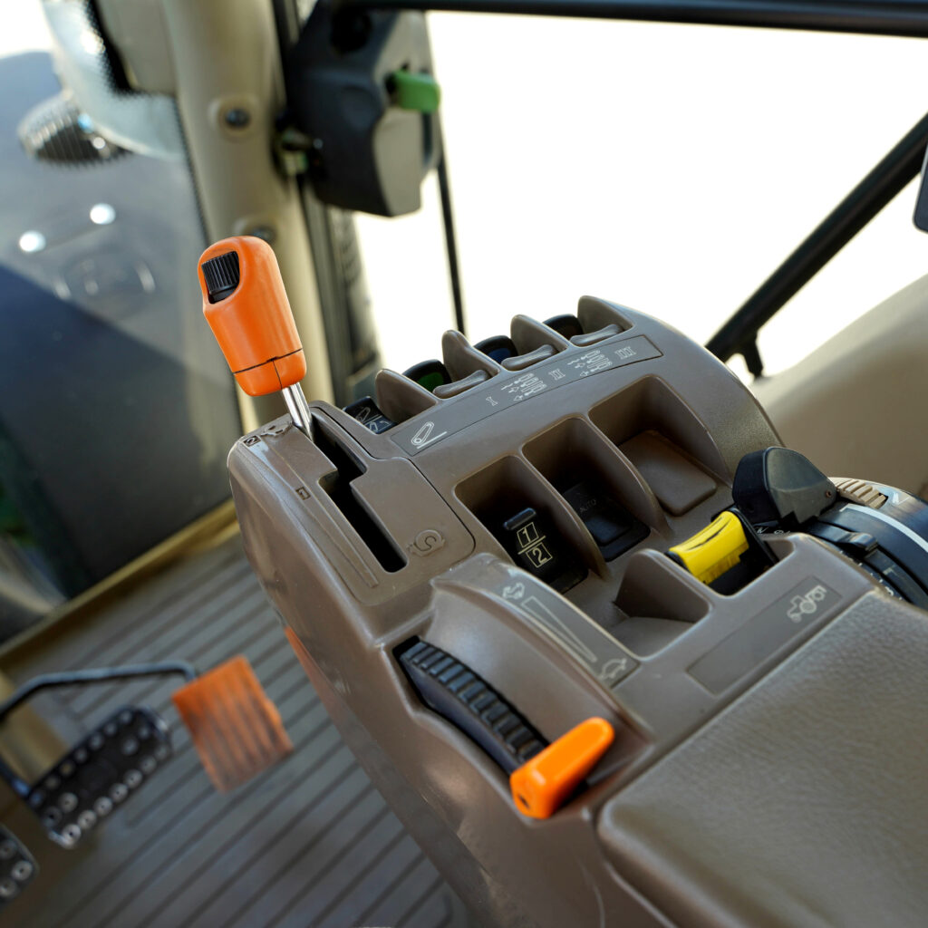 Blick in die Fahrerkabine eines Baumaschinengeräts mit orangefarbenem Joystick, mehreren Hebeln und markierten Bedienelementen in braunem Kunststoff.