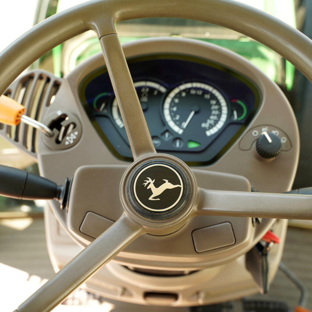 Durch das beige Lenkrad eines Traktors blickt man auf das Instrumentenpanel, während das markante Hirsch-Emblem zentral im hellen Fahrerkabinenlicht sitzt.