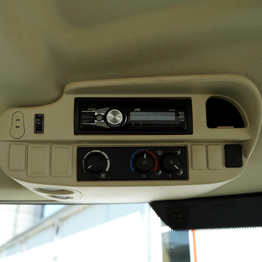 Beige Kabinendecke mit schwarzem JVC-Radio und Klimabedienung, dazu mehrere Schalter und ein gepunkteter Dachhimmel in einem Fahrzeug.