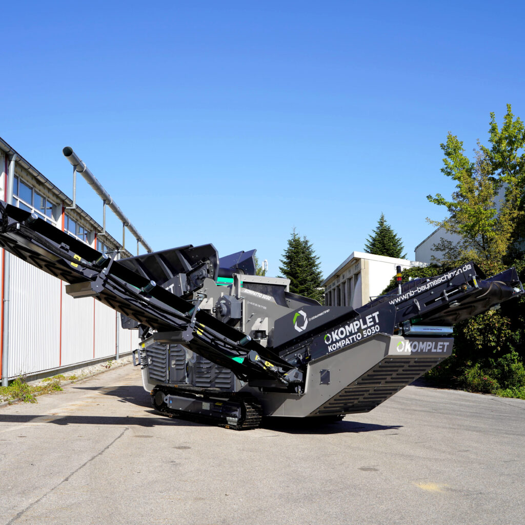 Große schwarze und graue Komplet KOMPATTO 5030 mit Förderbändern steht auf einem Firmengelände, daneben Hallen, Bäume und ein klarer blauer Himmel.