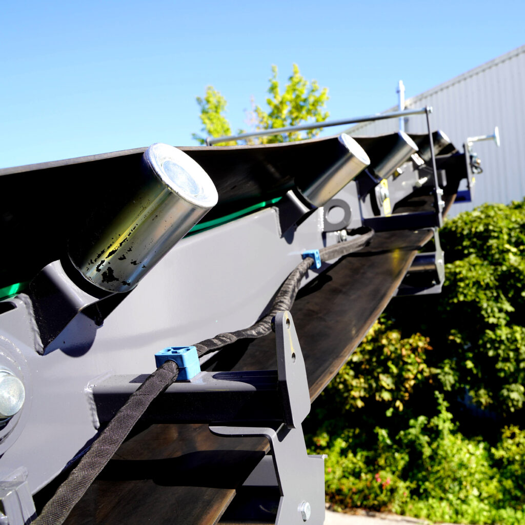 Nahaufnahme einer schwarzen Metallkonstruktion mit glänzenden Zylinderaufsätzen vor blauem Himmel und grünem Laub auf dem Werksgelände im Sonnenlicht.
