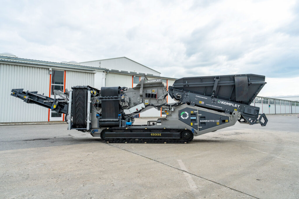 Ein grauer KOMPLET KOMPATTO 104 Brecher steht vor einer Industriehalle auf Beton, mit langem Förderband, Raupenfahrwerk und großem Trichter.