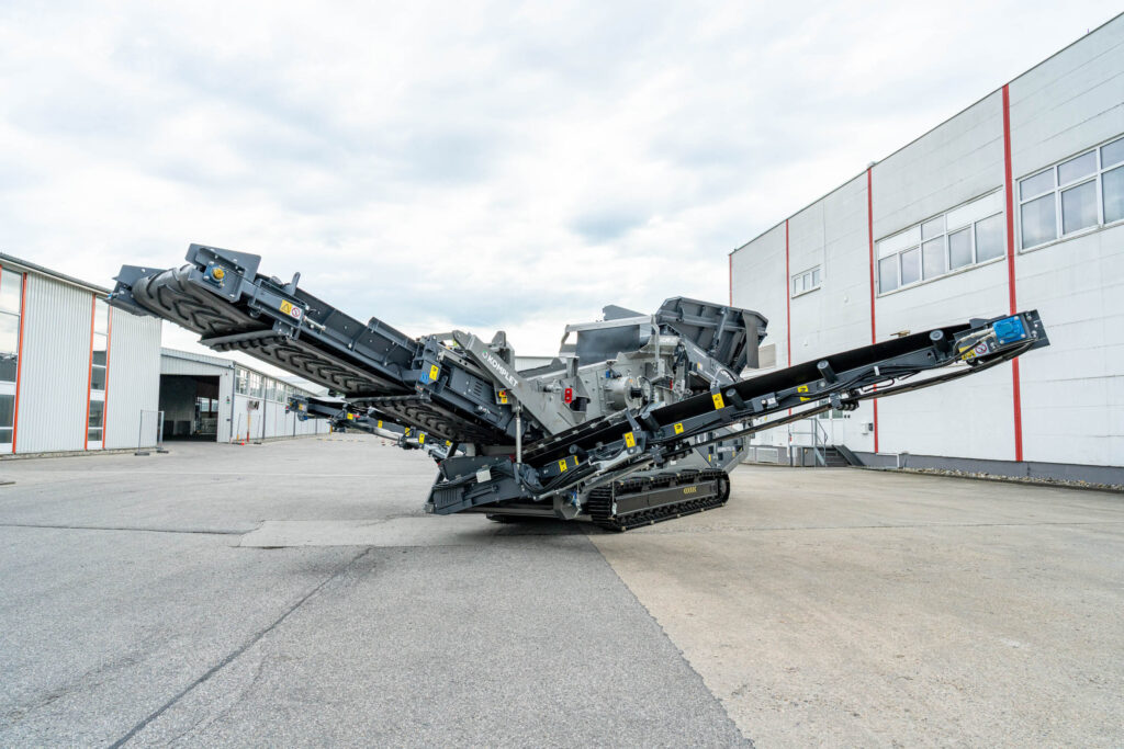 Ausgeklappte Komplet-Siebanlage in Grau auf einem Betonhof zwischen Industriegebäuden, mit langen Förderbändern und Kettenlaufwerk unter hellem Himmel.