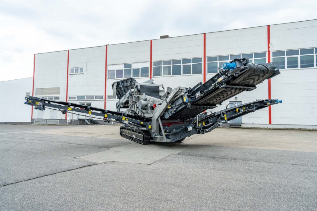 Eine schwarze und graue Recyclinganlage mit ausfahrbaren Förderbändern steht auf dem Hof vor einer weißen Industriehalle mit roten Streifen.