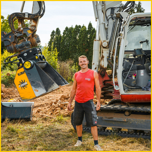 Ein Mann im roten T-Shirt steht vor einem Bagger mit Greifer, während links ein gelber Anbaukopf und ein Erdhaufen auf der Baustelle zu sehen sind.