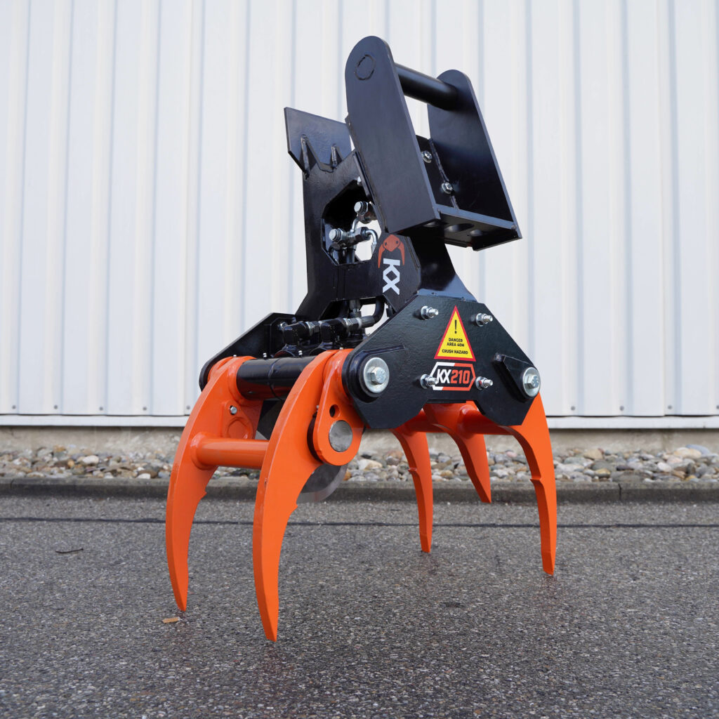 Schwarzer KX-210 Fällgreifer mit orangefarbenen Greifarmen und Hydraulikanschlüssen steht im Freien vor einer hellen Wellblechwand auf Asphalt.
