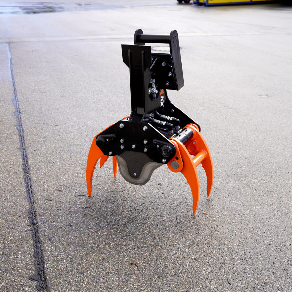 Orange-schwarzer Fällgreifer KX-210-4 mit vier Greifarmen und sichtbaren Hydraulikanschlüssen steht auf Asphalt vor hellem Hintergrund.