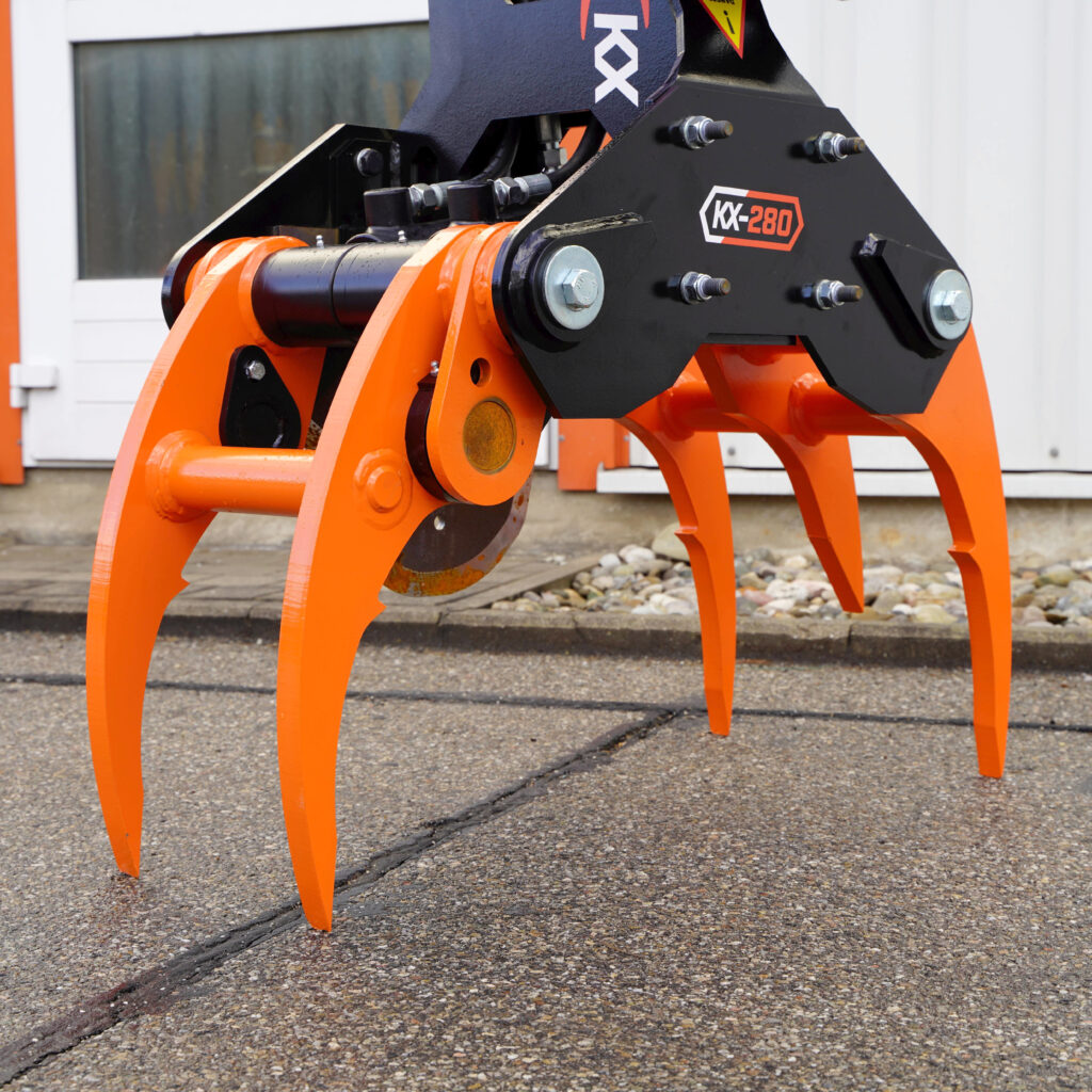 Ein orange-schwarzer KX-280-Fällgreifer mit vier gebogenen Zinken, sichtbaren Hydraulikschläuchen und massiven Bolzen steht vor einer hellen Wand auf Beton.