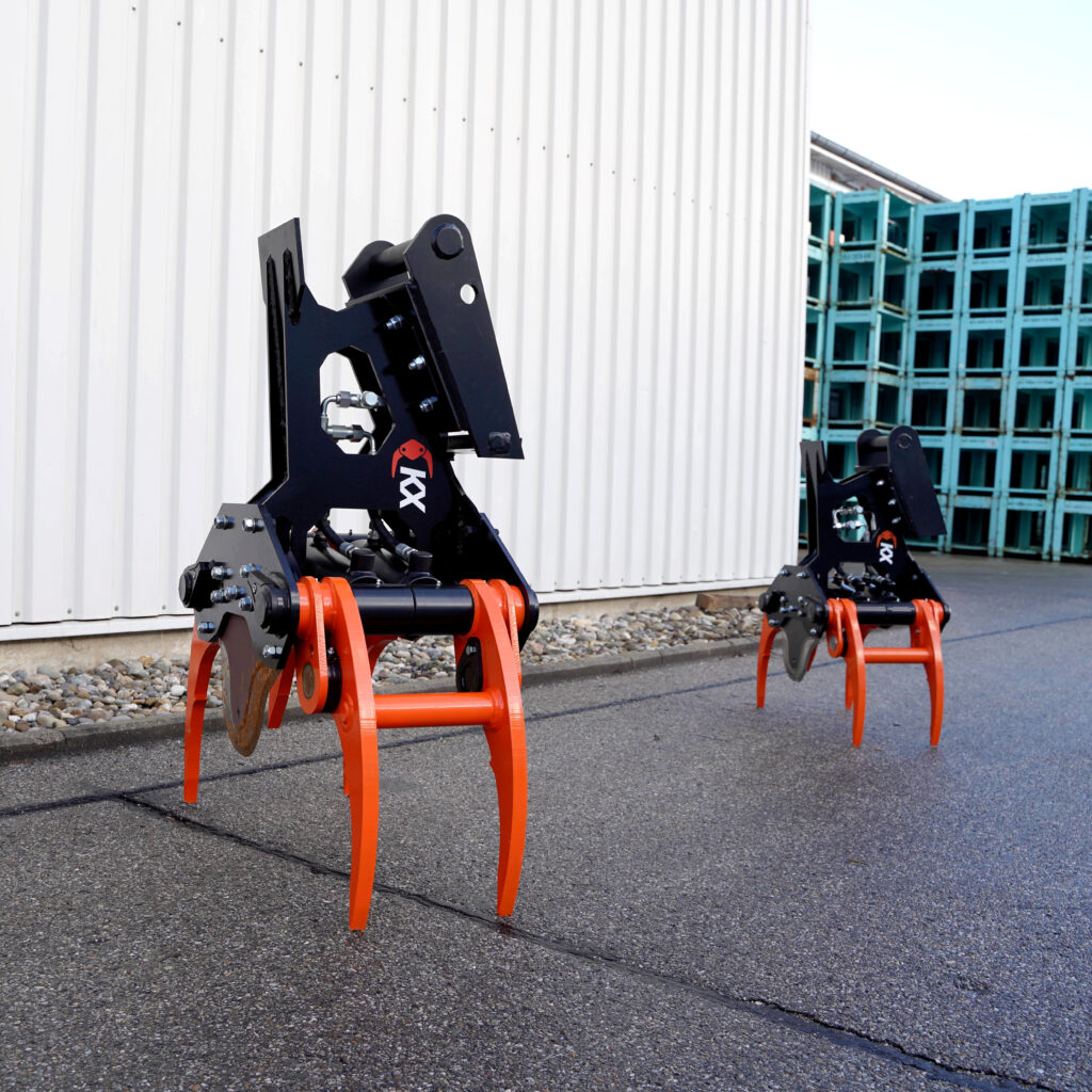 Zwei schwarze KX-280-4 Fällgreifer mit orangefarbenen Greifarmen stehen vor einer weißen Industriehalle auf nassem Asphalt neben blauem Stahlgestell.