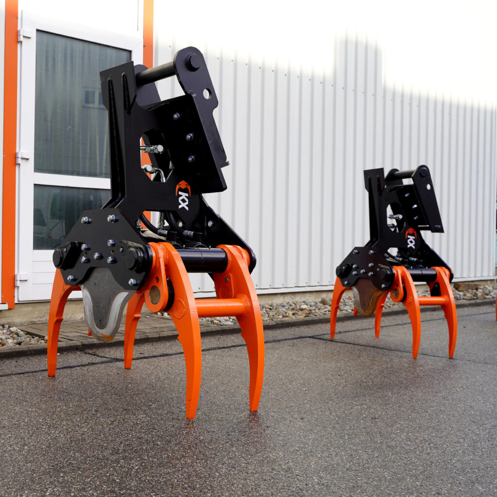 Zwei schwarze KX-350-4-Fällgreifer mit orangefarbenen Greifarmen stehen im Freien vor einer hellen Hallenwand auf nassem Asphalt bei NB Baumaschinen.