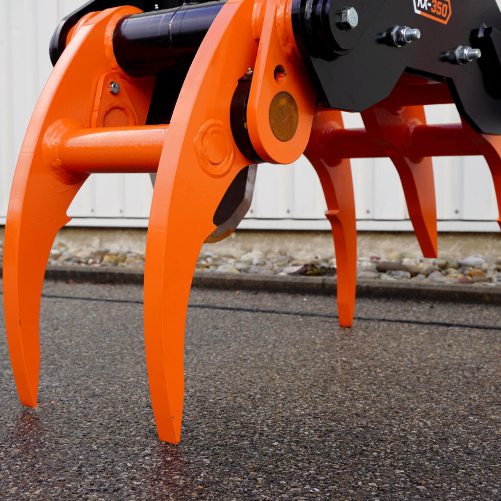 Ein orange-schwarzer Faellgreifer KX-350 mit massiven Greifarmen und Stahlbolzen steht in Nahaufnahme auf Asphalt vor einer hellen Wand.