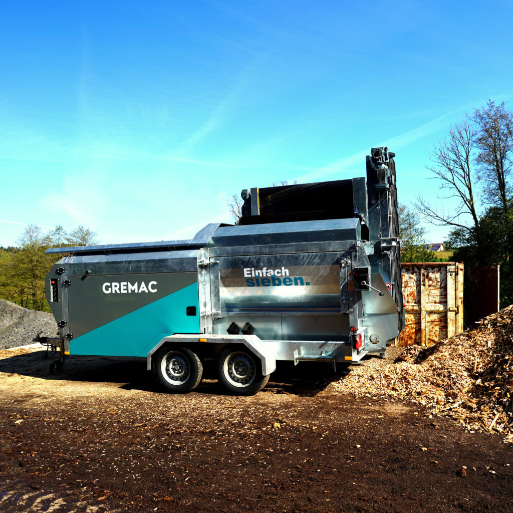 Ein turkis-silberner GREMAC-Radmobil-Siebtechnik-Anhänger steht auf einem Recyclinggelände zwischen Holzresten und Containern unter klarem Himmel.