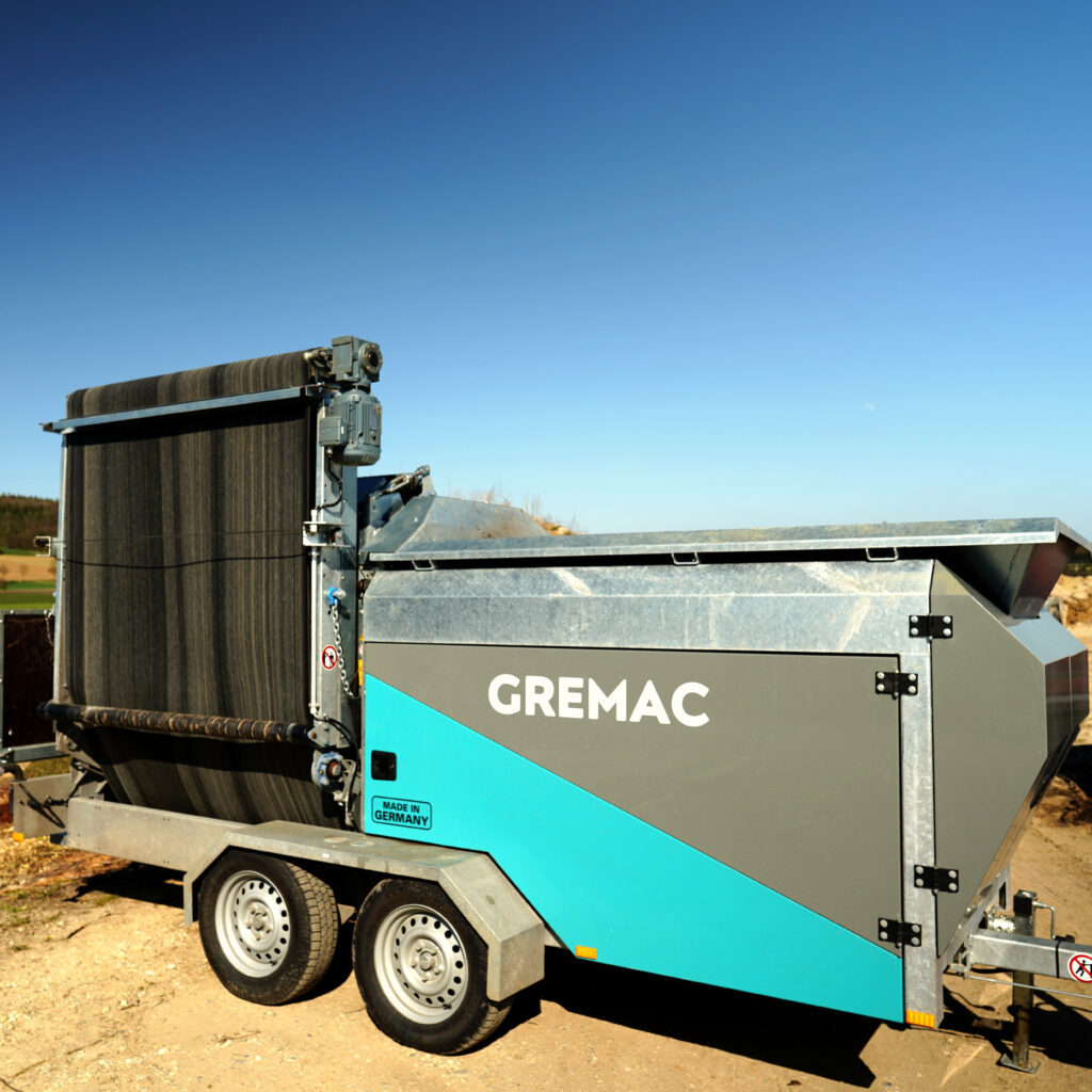 Ein mobiler GREMAC-Radmobil mit großem schwarzen Förderband, türkis-grauem Gehäuse und Tandemachsen steht auf einem sandigen Gelände unter blauem Himmel.