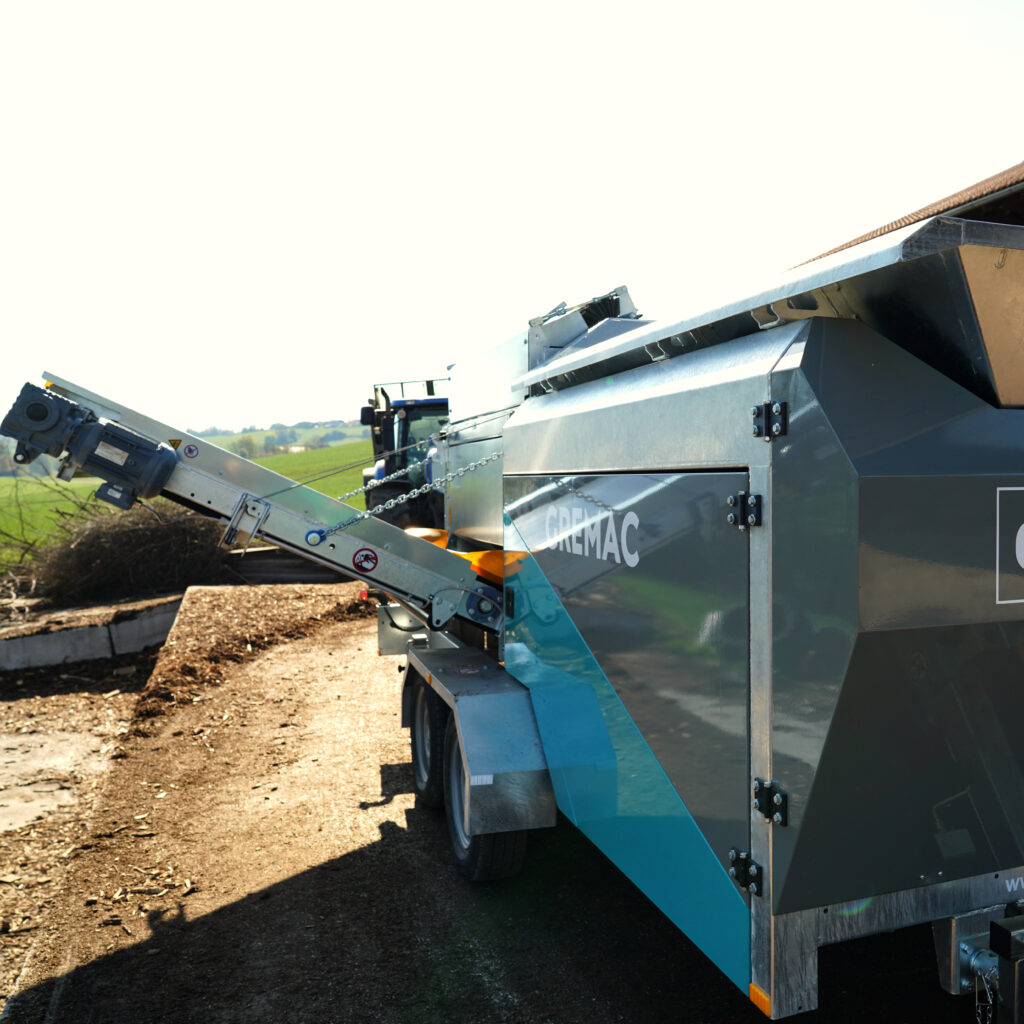 Ein GREMAC-Recyclinggerät mit Förderband steht auf einem Hof vor grünen Feldern, während die metallische Anlage im hellen Sonnenlicht glänzt.