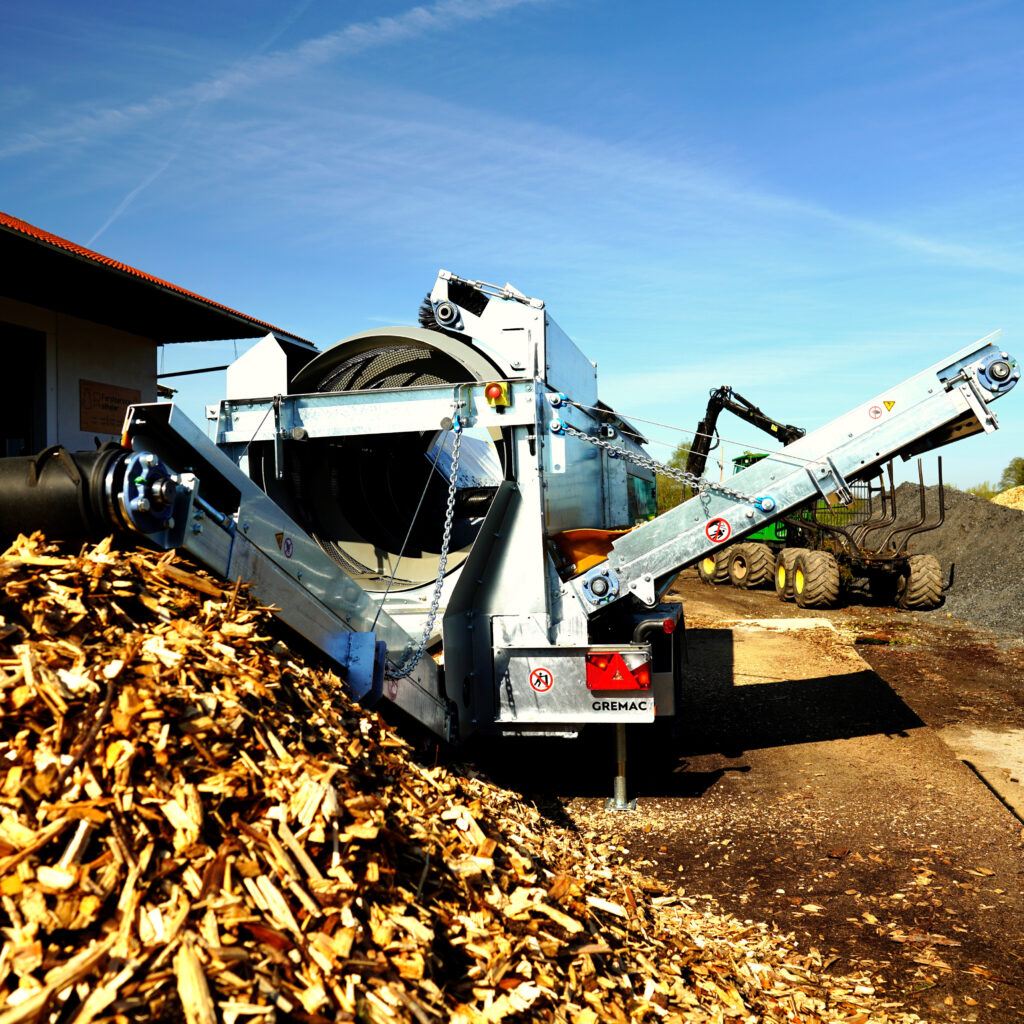 Eine GREMAC-Radmobilanlage verarbeitet Holzschnitzel, während Förderbänder, Trommel und Ketten im Freien unter blauem Himmel arbeiten.