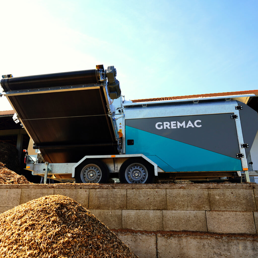 Ein GREMAC-Radmobil mit ausgefahrenem Förderband steht auf einem Recyclinghof, während unten ein großer Holzschnitzel-Haufen unter blauem Himmel liegt.