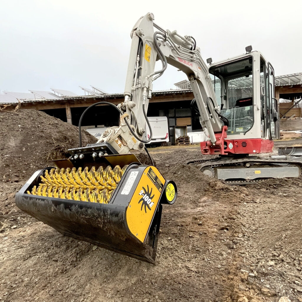 Ein weiß-roter Takeuchi-Raupenbagger arbeitet mit gelb-schwarzem GYRUstar 2-100E Sieblöffel auf einem schlammigen Recyclingplatz.