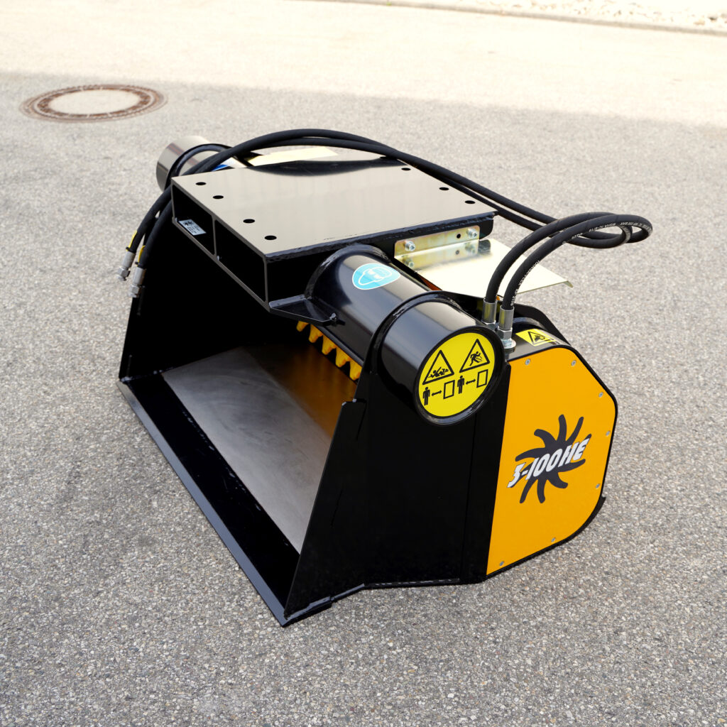 Ein schwarzer und gelber GYRUstar 3-100HE-5 mit Hydraulikschläuchen, Warnsymbolen und breiter Schaufel steht auf Asphalt vor hellem Hintergrund.