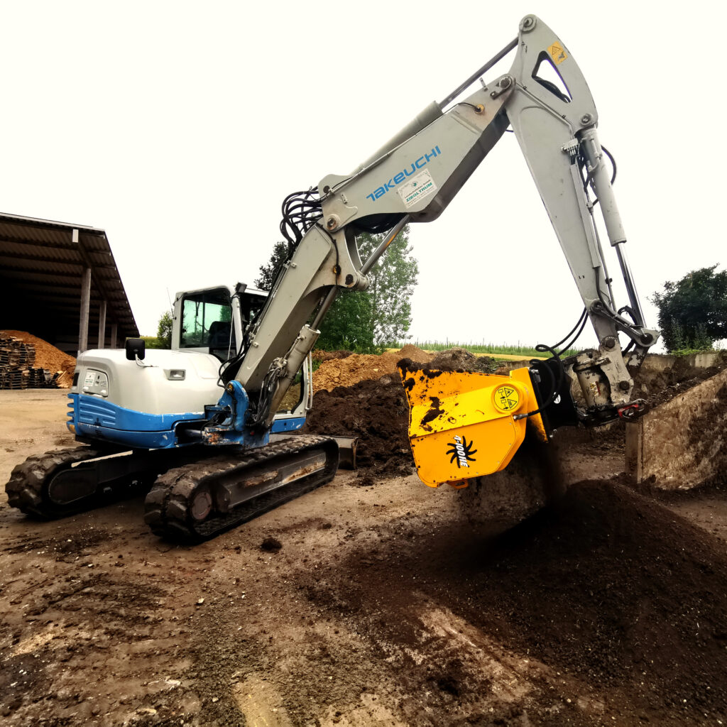 Ein weiß-blauer Takeuchi-Kettenbagger arbeitet an einer Erdgrube und drückt mit einem gelben Anbaugerät Material neben einem Schuppen auf dem Hof.