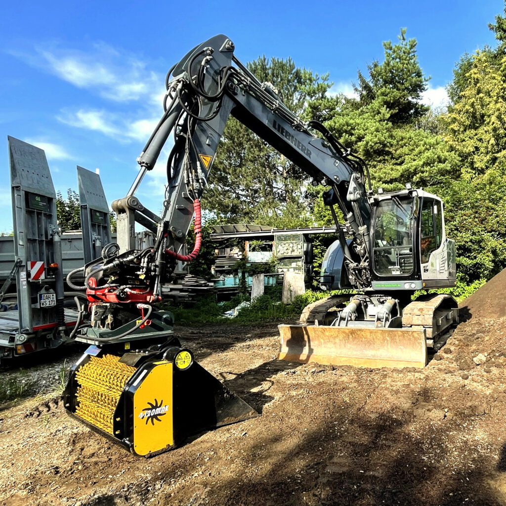 Ein Liebherr-Bagger steht auf einem Waldgelände neben einem Anhänger, während vorne ein gelb-schwarzes GYRUstar-Anbaugerät im Boden ruht.
