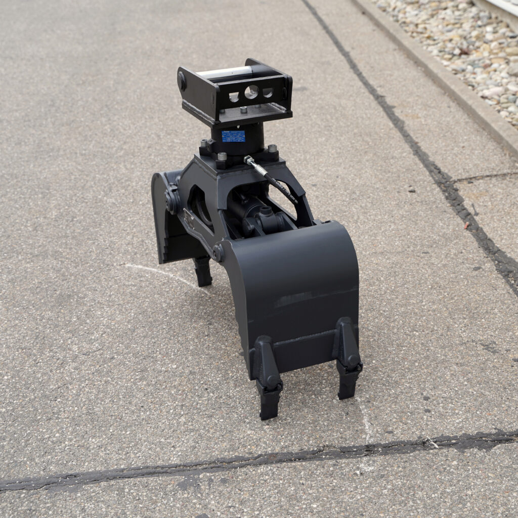Schwarzer hydraulischer Greifer mit massiver Stahlkonstruktion und drei Zinken steht auf Asphalt neben einem Bordstein und zeigt Schläuche sowie Anschlüsse.