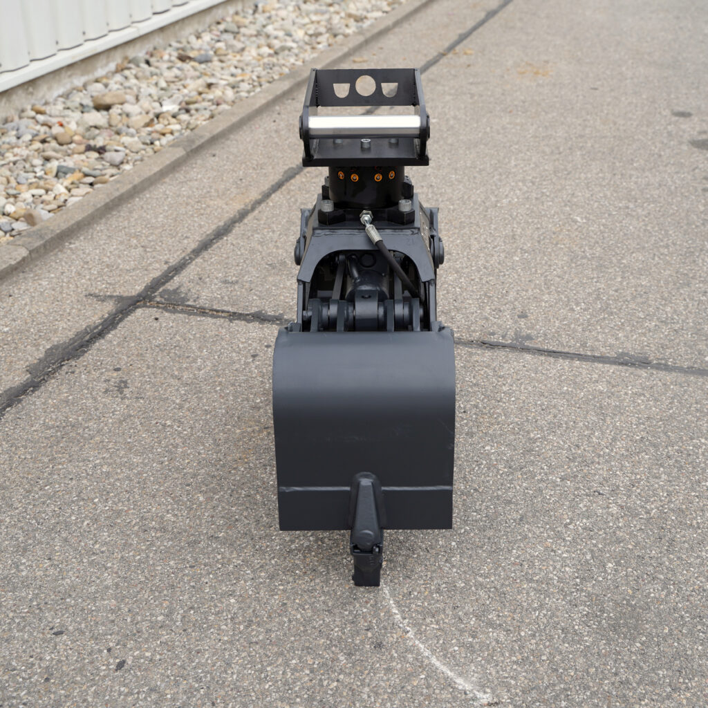 Ein schwarzer Greifer mit Hydraulikanschluss steht auf Asphalt, davor ist ein breiter Greiflöffel zu sehen; links verläuft ein Kiesstreifen am Rand.
