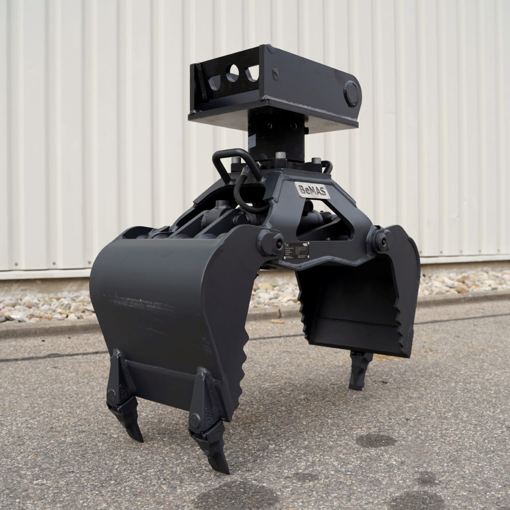 Der schwarze BeMAS-Greifer mit massiven Stahlbacken und oberer Aufnahme steht im Freien vor einer hellen Metallwand auf Asphalt.