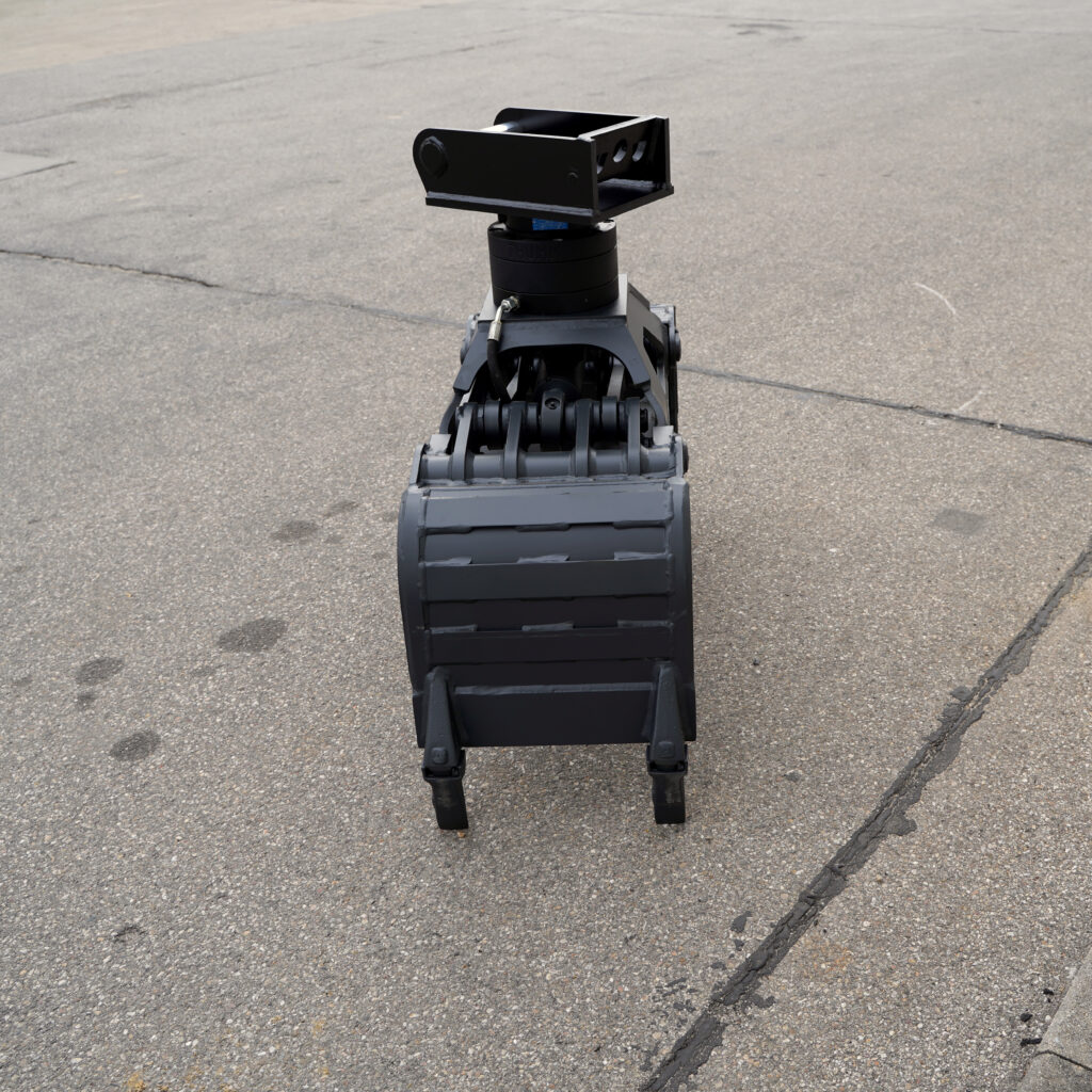 Ein schwarzer Baggergreifer mit breiter Aufnahme und massiver Mechanik steht auf grauem Asphalt, die Blickrichtung zeigt frontal auf die schwere Konstruktion.