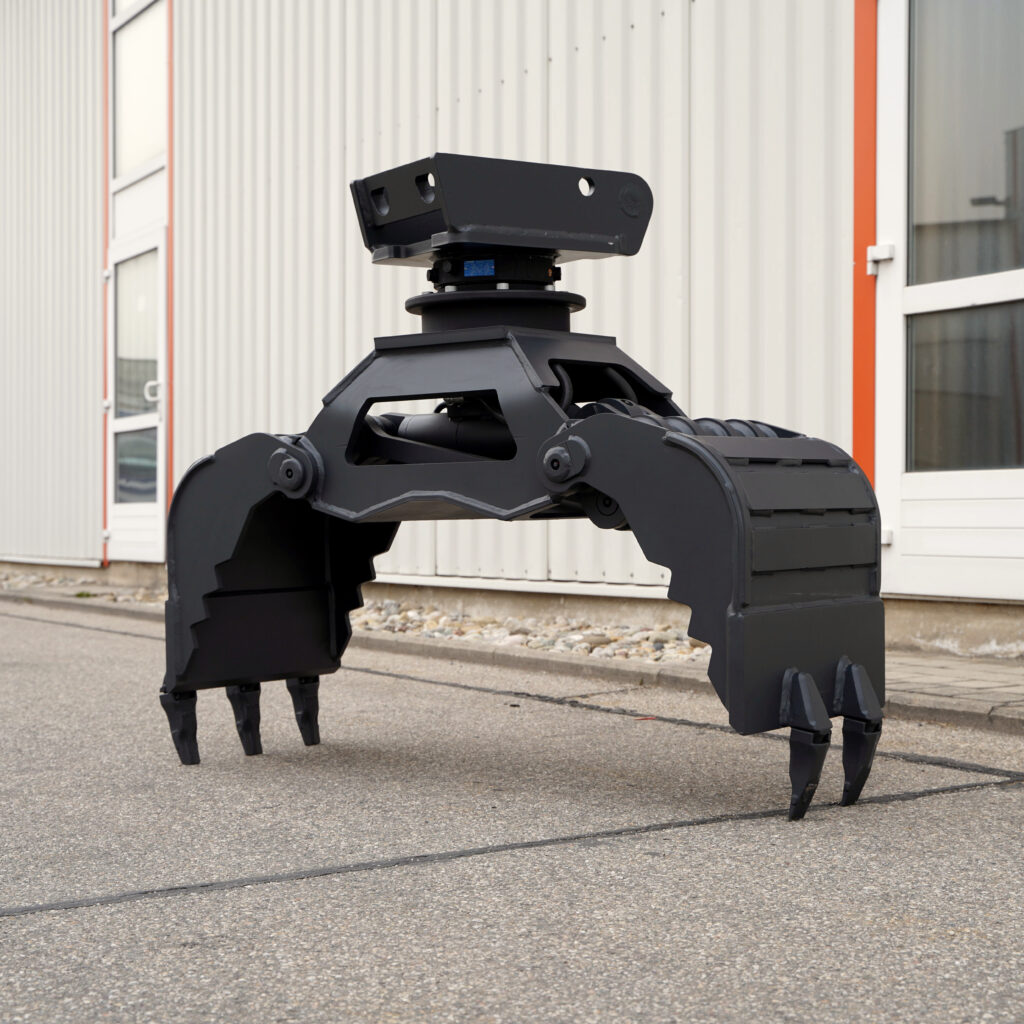 Ein schwarzer hydraulischer Greifer mit vier klauenartigen Zinken steht vor einer hellen Industriehalle auf dem Asphalt neben Fenstern und roten Streifen.
