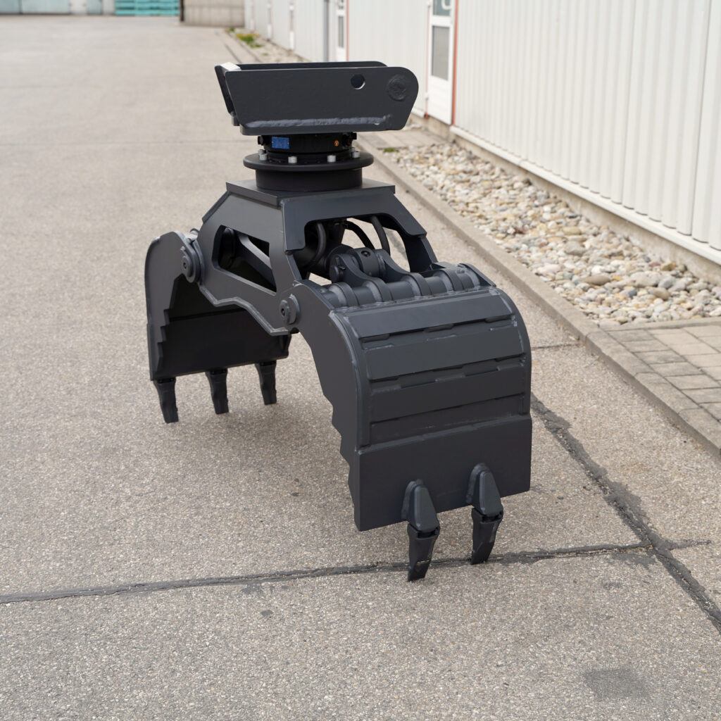 Schwarzer Greifer für Bagger mit vier Zinken und Drehkopf steht auf Asphalt vor einer hellen Industriehalle und zeigt massive Stahlkonstruktion.