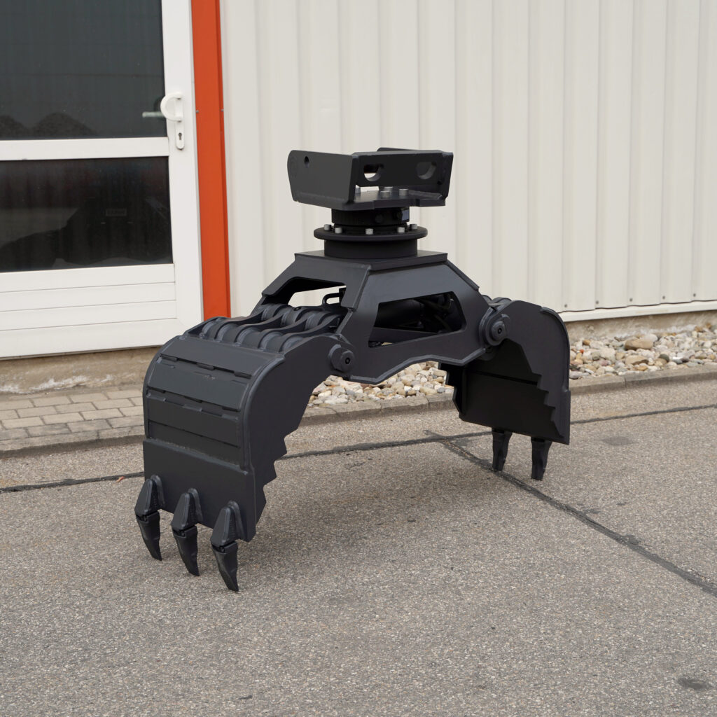 Ein schwarzer Baggergreifer mit Drehkopf und mehreren Zähnen steht vor einer hellen Hallenwand auf dem Asphalt im Freien.