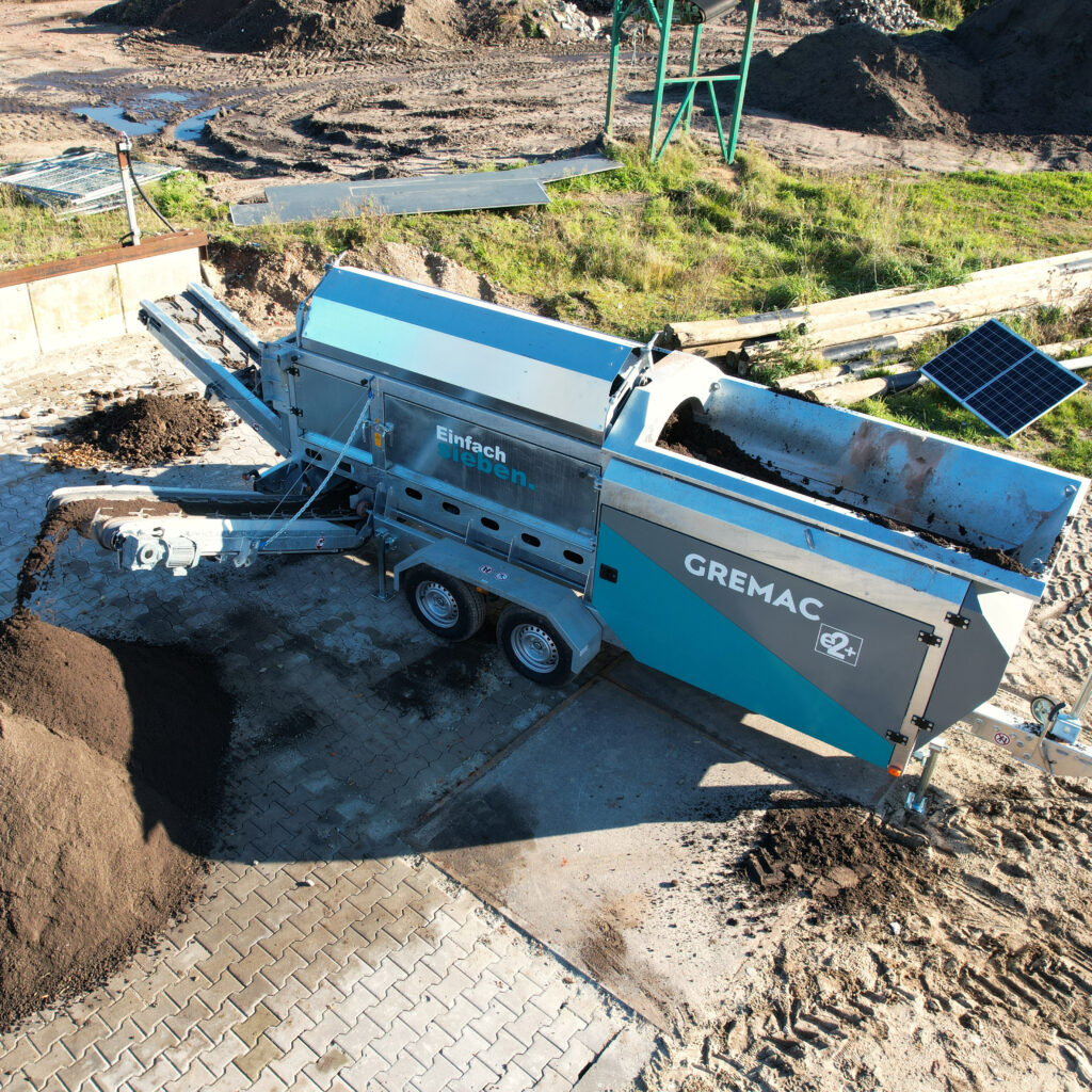 Ein Gremac e2+ Siebmaschinenanhänger fördert dunkles Erdmaterial auf einer Baustelle, umgeben von Sandhaufen, Pflaster und grüner Wiese.