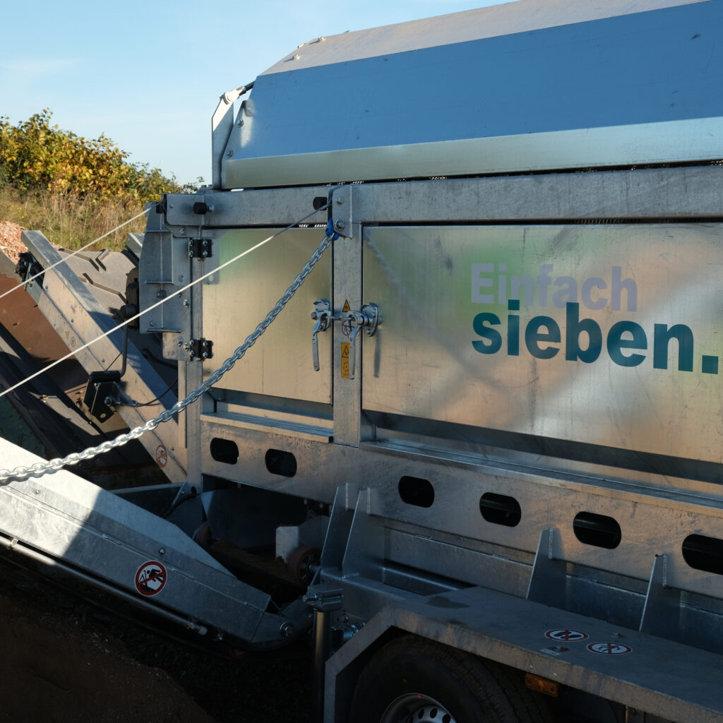 Ein silberner Gremac-Siebaufbau mit großen Metallpanelen, Ketten und Verstrebungen steht im Freien vor blauem Himmel und Büschen.