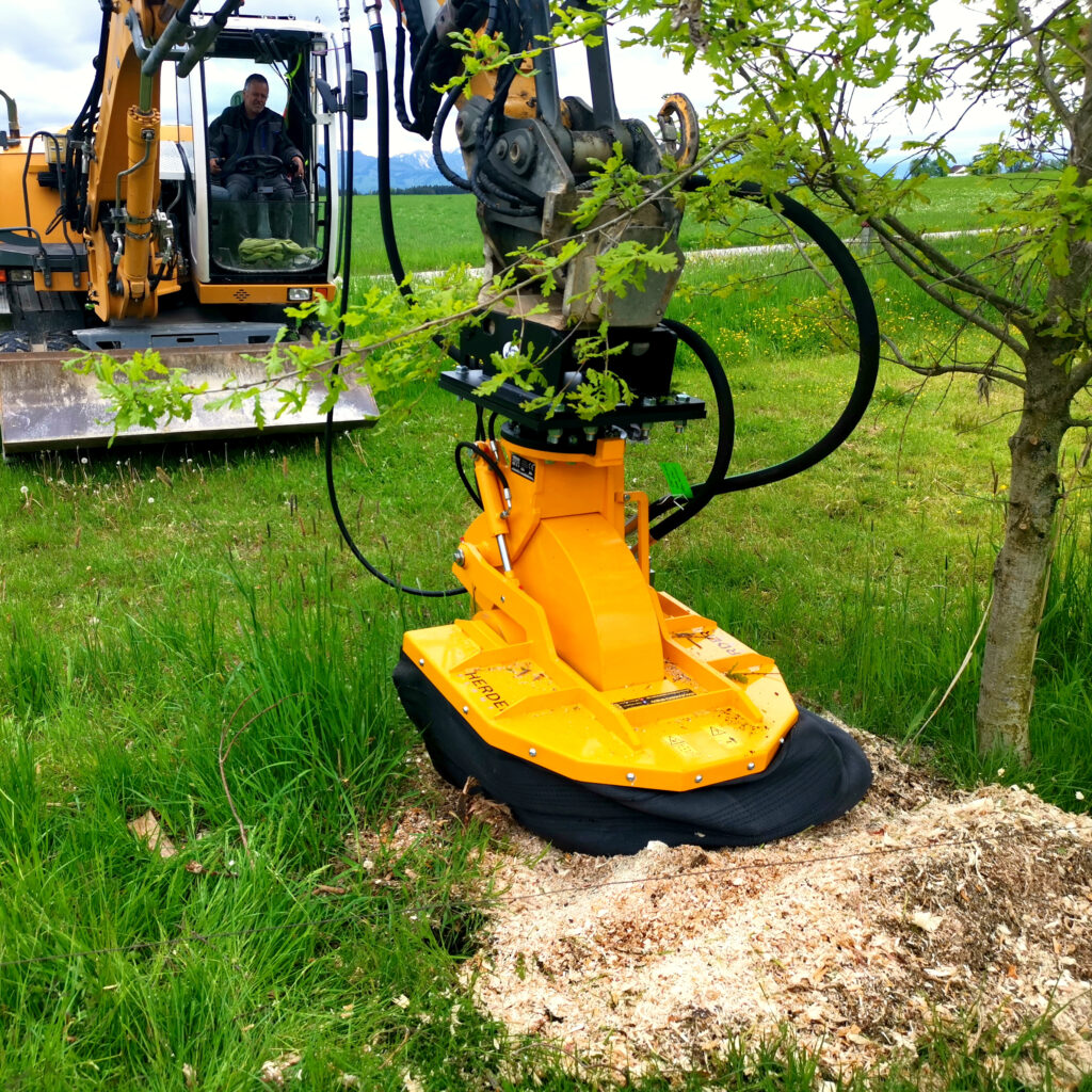 Ein gelber Herder Stubbenfräse SCE-630H-2 arbeitet an einem Baumstumpf neben einem Traktor, während frische Grünfläche und Äste die Szene rahmen.