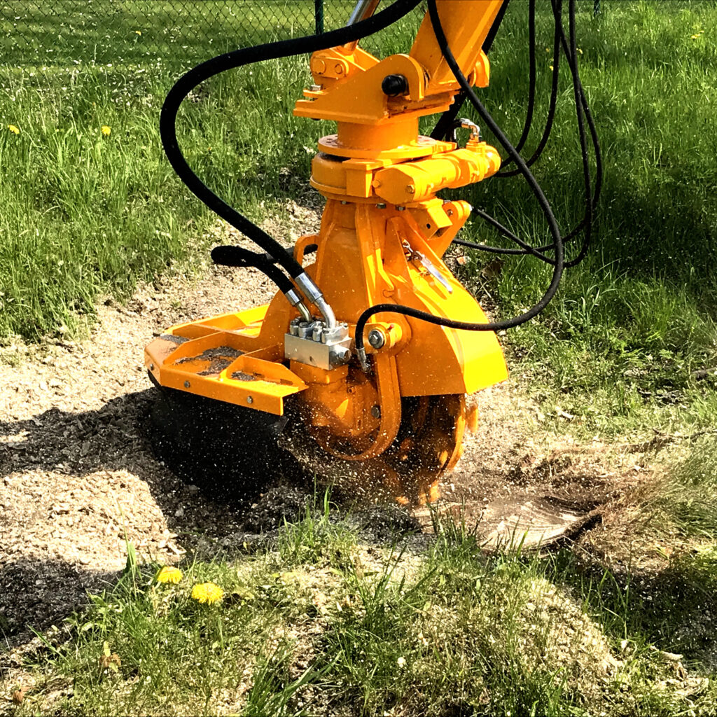 Der orange Herder Stubbenfräse SCE-630H-3 fräst einen Baumstumpf im Gras, während Holzspäne und schwarze Hydraulikschläuche sichtbar sind.