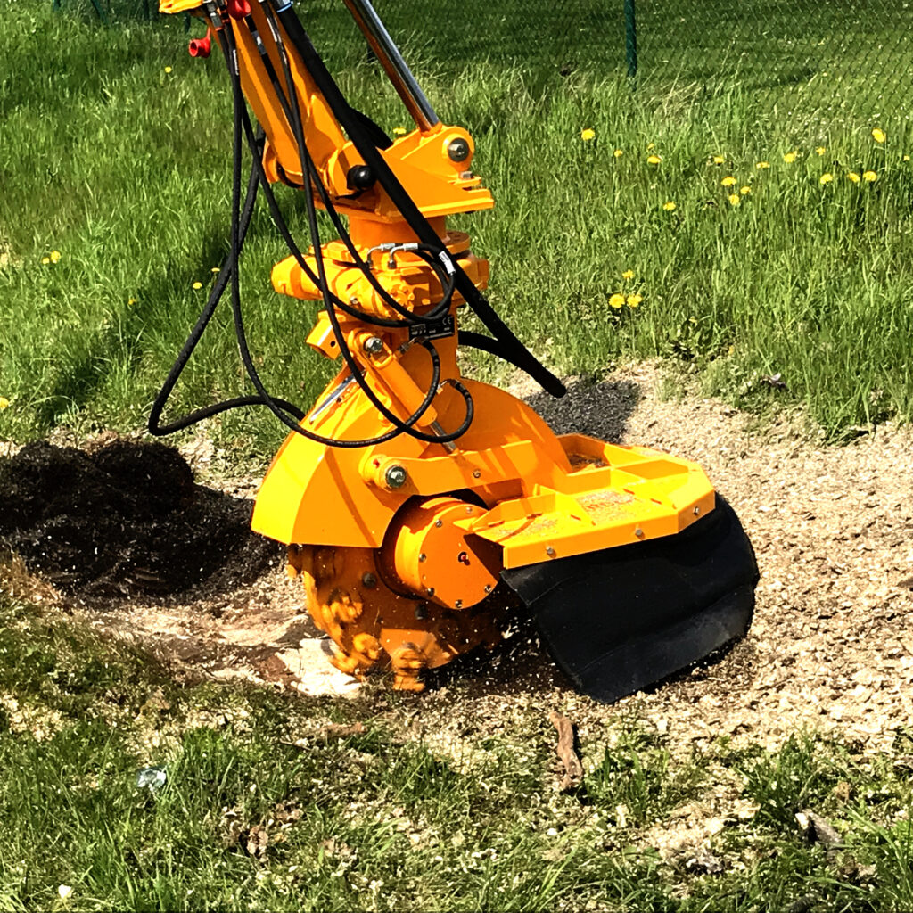 Ein orangefarbener Herder-Stubbenfräse arbeitet im Gras und fräst einen Baumstumpf neben einem Kiesstreifen, während schwarze Schläuche mitlaufen.