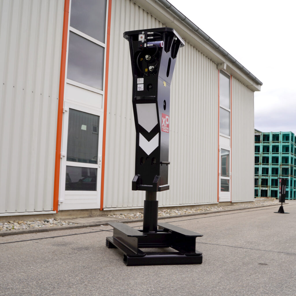 Schwarzer Hydraulikhammer XP1500 mit sichtbaren Anschlüssen und Schutzplatte steht vor einer hellen Industriefassade mit orange umrahmten Fenstern.