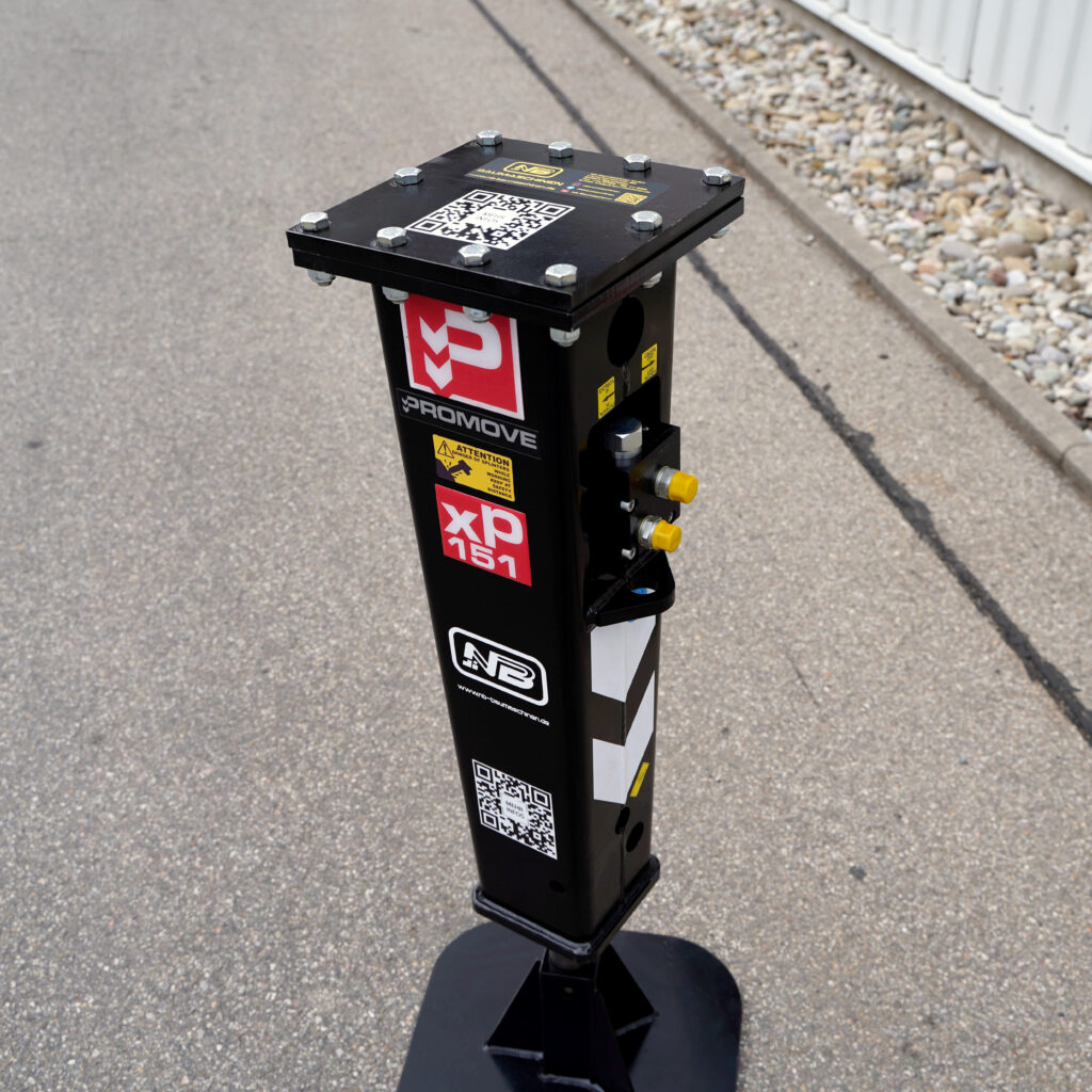 Schwarzer Hydraulikhammer mit PROMOVE- und NB-Aufklebern, roter XP151-Markierung, gelben Anschlüssen und QR-Codes steht auf Asphalt neben einem Gebäudekiesstreifen.